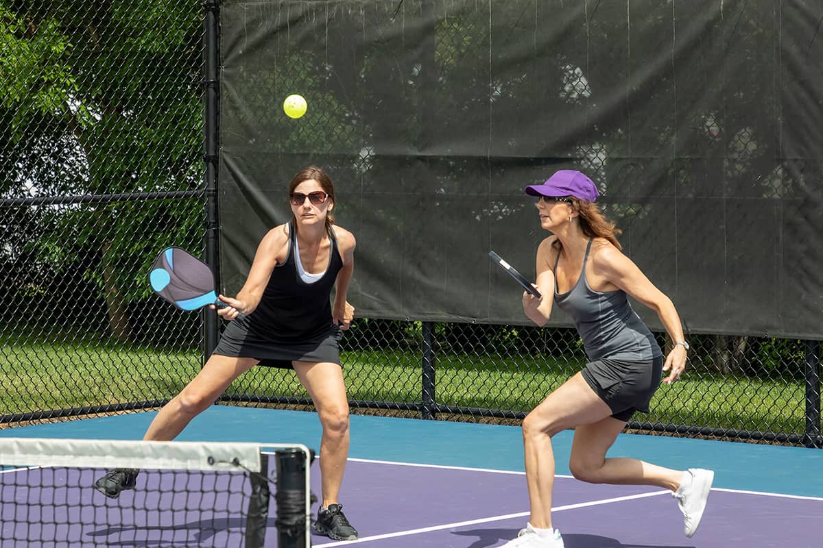 Two women run towards the net during a match of tennis on the NJAC courts