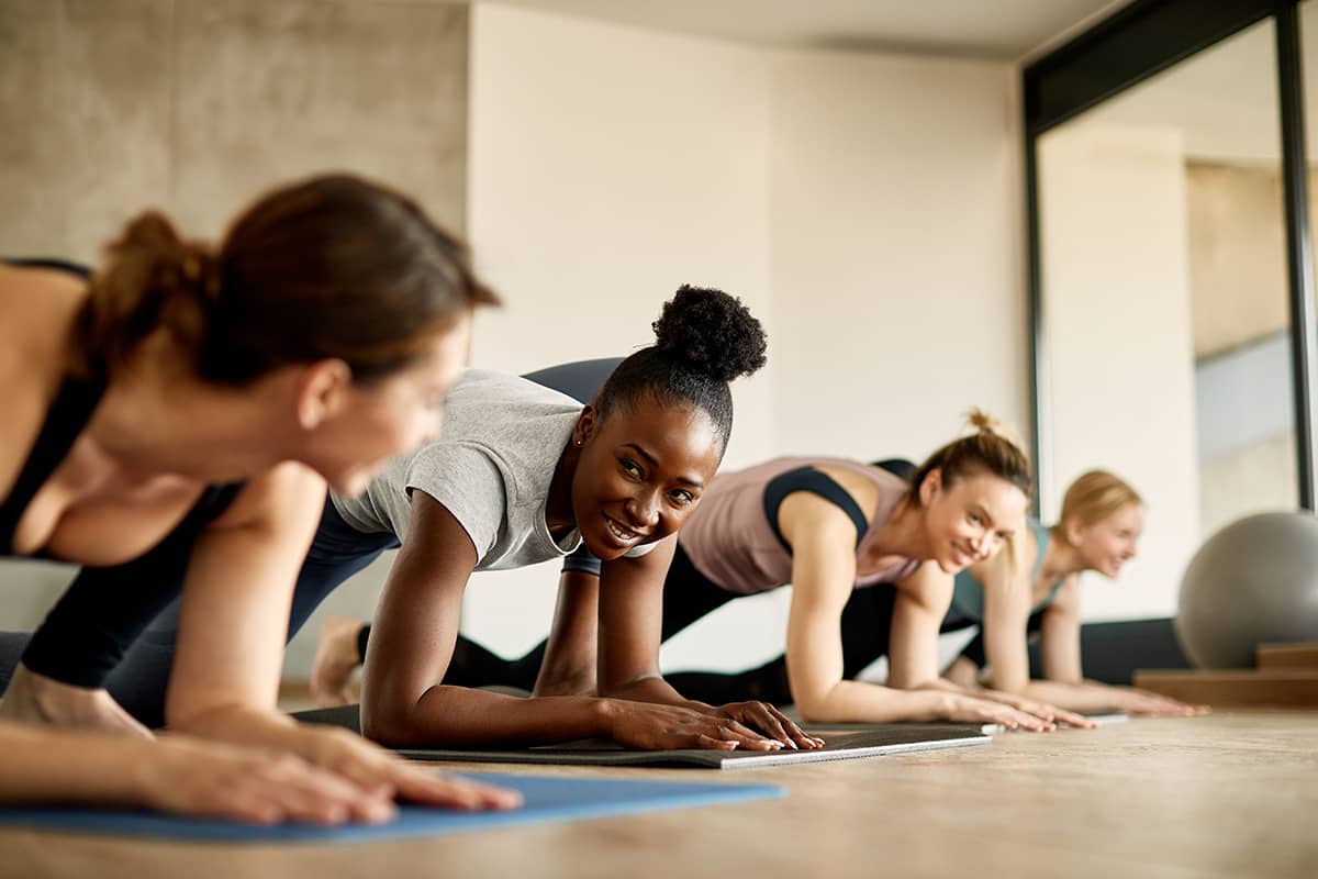 Women on yoga mats hold planks and smile at each other during a group fitness class